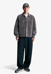 Szara kurtka sztruksowa z dwoma dużymi przednimi kieszeniami, zapięciem na guziki i luźnym krojem, zestawiona z luźnymi granatowymi spodniami i sportowymi butami.