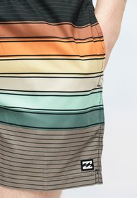 Ενήλικας που φοράει πολύχρωμα οριζόντιαριγέ σορτς με το χέρι στην τσέπη, δείχνοντας το ραμμένο στρίφωμα και μικρό μαύρο λογότυπο.