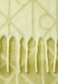 Couverture jaune clair avec une surface douce et texturée, présentant un motif géométrique et des bords frangés. Le matériau semble ressemblant à de la laine et est confortable.