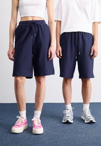 Shorts de survêtement bleu marine en tissu texturé, avec taille élastique à cordon, fentes sur les côtés, portés avec des baskets colorées et des chaussettes de sport blanches.