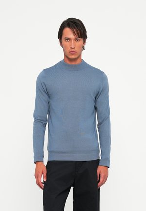 Man wearing a fitted blue long-sleeve sweater with mock neck and black pants, standing against a plain white background.