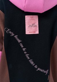 Czarna, brązowa i różowa bluza z kapturem. Posiada widoczną metkę i różowy napis w kursywie: "Każdy oddech może być listem miłosnym do samego siebie."