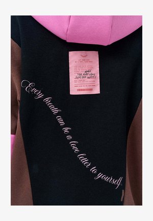 Mikina s kapucí v černé, hnědé a růžové barvě. Obsahuje viditelnou nálepku a růžový nápis psaný kurzívou: "Každý dech může být milostním dopisem pro tebe samého."