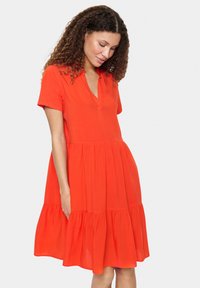 Rochie roșie, cu mâneci scurte, de tip V, fustă în straturi și croială lejeră. Fabricată dintr-un material ușor, cu o textură netedă.