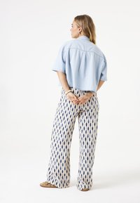 Camisa corta de color azul claro con mangas cortas combinada con pantalones beige de estampado que presentan diseños de gotas de agua azules. Modelo de espaldas.