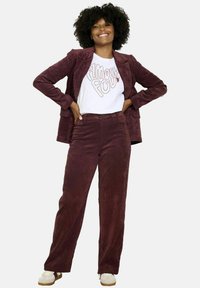 Donna sorridente con capelli ricci, che indossa un blazer e pantaloni in corda color bordeaux, una maglietta bianca con grafica e sneaker color beige, in piedi con le mani sui fianchi.