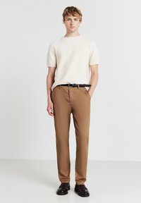 Maglione beige a maniche corte con texture abbinato a pantaloni marroni su misura e una cintura nera. Il modello indossa scarpe slip-on scure. Design semplice e moderno.