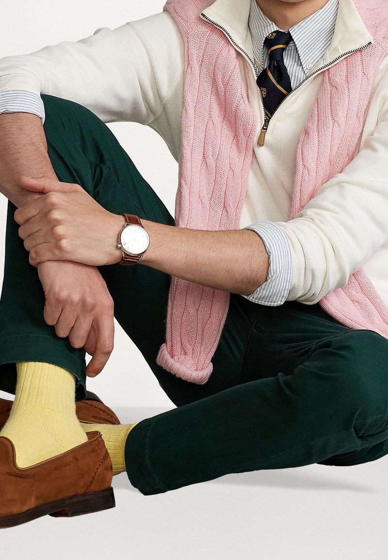 Personne assise en tailleur portant un pantalon vert, des chaussettes jaunes, des chaussures marron, un pull blanc, un gilet en tricot rose, une chemise rayée, une cravate noire et une montre-bracelet.