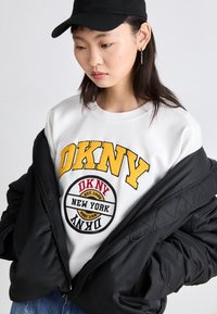 Λευκή μπλούζα με κίτρινο και μαύρο λογότυπο "DKNY", φορεμένη κάτω από μια μαύρη επενδεδυμένη ζακέτα. Το μοντέλο φοράει ένα μαύρο καπέλο με υφή.