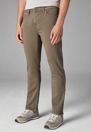 Uske uskom slim fit, maslinasto zelene traperice s glatkom teksturom, dizajnom s pet džepova i kontrastnim šavovima. Vidljive su siva sportska obuća.