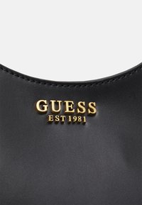 Sac à main synthétique noir avec une texture lisse et un logo doré "GUESS EST 1981". Le bord courbé souligne son design moderne.