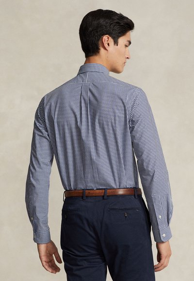 Mann in marineblau-weiß kariertem Langarmhemd, das in marineblaue Hose mit braunem Gürtel gesteckt ist, von hinten gesehen.