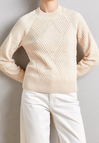 Person som bär en gräddfärgad strukturerad stickad tröja med geometriskt ribbat mönster och vita byxor, står med händerna bakom ryggen.