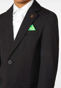 Laps, kes kannab musta bleiserit rohelise tasku ruuduga ja valget lipsu, millel on õlavarrelt oranž õmblusrist.