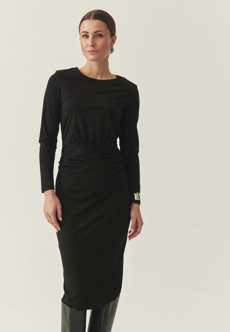 Robe noire à manches longues avec taille froncée, silhouette ajustée et longueur midi. Portée avec un bracelet argenté brillant et des bottes montantes jusqu'aux genoux.