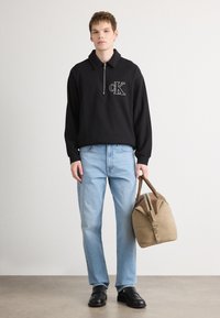 Must-haves must-olla mustra-ärri moosybyn 'CK' logo, koos heledate siniste teksade ja mustade kingadega, kandes pruuni reisikotti.