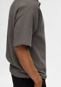 Chemise en tricot gris texturé à manches courtes avec une coupe décontractée et un col à bouton. Le tissu semble doux et respirant, adapté pour un usage décontracté.