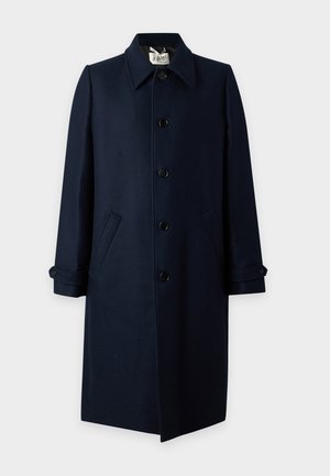 Jeune homme debout portant un manteau bleu marine foncé, une chemise rayée, un pantalon beige et des baskets beiges avec des lacets roses sur un fond blanc uni.
