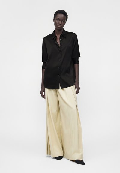 Γυναίκα με φαρδιά μαύρη πουκαμίσα με κουμπιά και μπεζ παντελόνες με φαρδιά πόδια στέκεται μπροστά σε απλό λευκό φόντο.