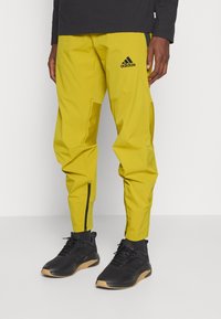 Gelbe Sporthose mit schmal zulaufender Passform, ausgestattet mit Reißverschlüssen an den Knöchelabschlüssen, strukturierten Einsätzen und einem schwarzen adidas-Logo am oberen Bein.