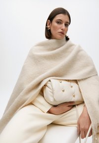 Kremasti vunski plašt nanesen preko svijetle kremaste odjeće, uparen s teksturiranom kremastom torbom koja ima zlatne metalne detalje.