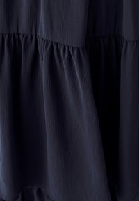 Temno modra večplastna obleka z teksturirano tkanino. Ima nabrano zavezovanje v pasu in valovit silhueto.