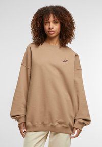 Pruun ülekaaluline sweatshirt, millel on ümmargune kaelus, langevad õlad ja soonikkoes mansetid. Vasakul rinnal on väike logo.