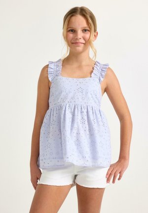 Pige i en ærmeløs lyseblå bluse med hulmønster og flæse-stropper samt hvide shorts, stående med den ene hånd på hoften foran en ensfarvet baggrund.