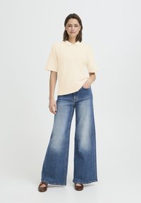 Femme se tenant debout, une main dans la poche, portant un haut en tricot beige à manches courtes, un jean bleu à jambes larges et des chaussures brunes, sur fond blanc.