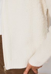 Pullover bianco testurizzato che presenta una combinazione di modelli di maglia liscia e soffice, con un dettaglio con zip e polsini a coste.