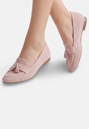 Femme portant des mocassins rose clair perforés avec des pompons, montrée de mi-mollet vers le bas sur un fond blanc uni.