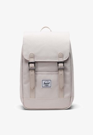 Mochila de un beige claro con solapa, dos hebillas en la parte frontal y un parche con logo. Hecha de un tejido duradero, con forma rectangular y textura suave.