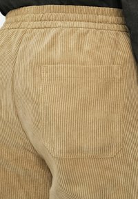 Béžové kordové nohavice so širokým elastickým pásom a jednou zadnou vreckom. Tkanina má vertikálne ribbované textúry pre pridanú detail.