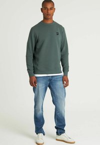 Felpa verde con texture, colletto a girocollo e un piccolo logo sul petto, abbinata a jeans azzurri chiari e sneakers bianche.