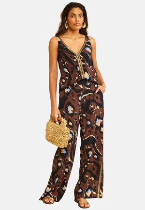 Kobieta ubrana w brązowy, czarny i niebieski top bez rękawów w zwierzęce wzory i spodnie, trzyma teksturowaną beżową torebkę i nosi złote sandały.