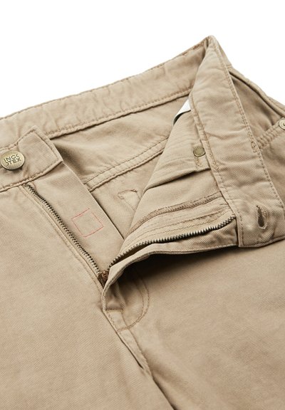 Pantalones de algodón beige con cremallera, cierre de botón y un sutil detalle de costura roja en la cinturilla. Textura suave y corte clásico.