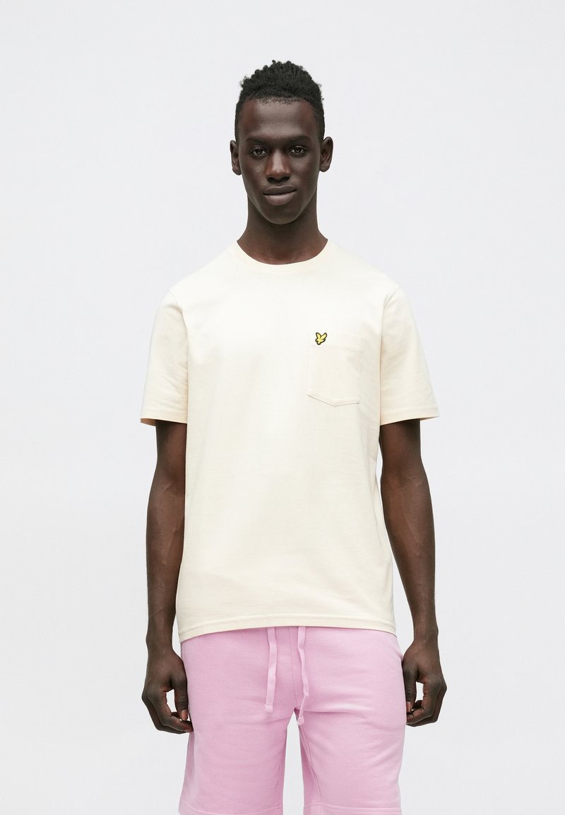 Kremfarvet T-shirt med korte ærmer, brystlomme og gult sommerfuglelogo. Kombineret med pink shorts, vist mod en neutral baggrund.