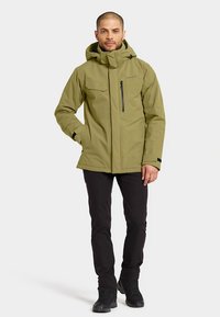 Olivengrøn vandtæt jakke med tætsiddende design, lynlåslommer, justerbar hætte og sorte detaljer. Båret med sorte bukser og støvler.