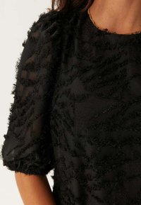 Top nero con texture, realizzato in morbido tessuto trasparente con dettagli a motivo in rilievo. Maniche corte a sbuffo che aggiungono volume al design.