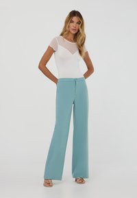 Un top bianco aderente con maniche corte trasparenti abbinato a pantaloni larghi color teal chiaro con chiusura a bottone e texture liscia.