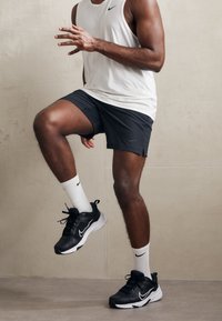 Baskets de sport noires Nike avec semelles blanches, shorts gris et débardeur léger. Chaussettes blanches avec un logo swoosh noir. Modèle dans une pose dynamique.