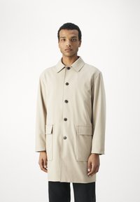 Beige trench coat klasszikus gallérral, elöl gombos záródással és két nagy foltpockkal. Simaszerkezet és középhosszú dizájn.