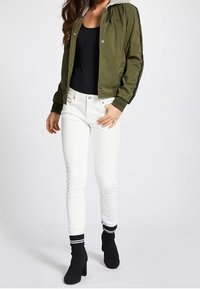 Olivgrön bomberjacka med grå huva, svarta sidosträck och tryckknappar, kombinerad med vita åtsittande jeans och svarta klackstrumpor.