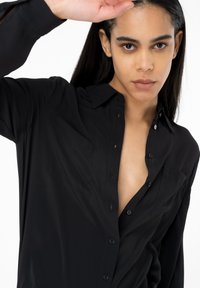 Camicia nera con colletto a punta, due tasche sul petto e una texture liscia, caratterizzata da una vestibilità comoda e un collo aperto.