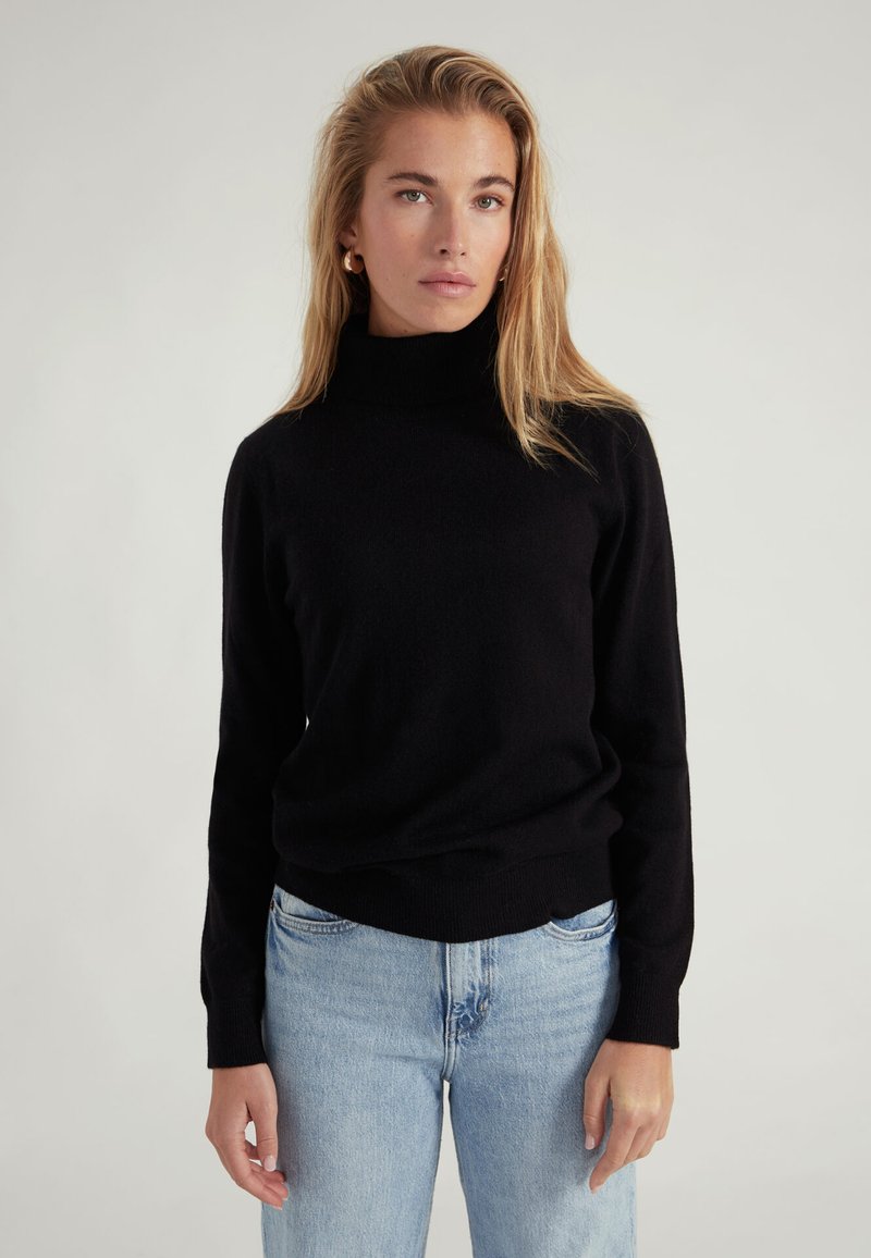 Maglione a collo alto nero realizzato in tessuto morbido, dal design aderente con polsini e orlo a costine, indossato con jeans azzurri chiari.