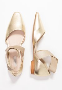 Bailarinas metálicas doradas con punta cuadrada, lados abiertos y correas de satén cruzadas. Presentan un bajo tacón de madera y una plantilla suave en tono beige.