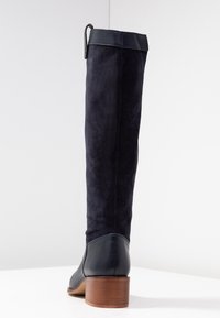 Botki do kolan w ciemnonavyj kolorze z zamszu, z gładkim skórzanym noskiem i drewnianym obcasem w kształcie bloku. Posiadają skórzaną taśmę na górze.
