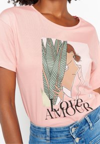 T-shirt di cotone rosa con una grafica che raffigura una donna con una pianta e la scritta "Love" in lettere nere in grassetto. Maniche corte, scollo rotondo.