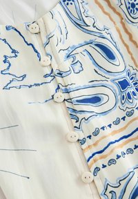 Gros plan sur un tissu crème avec des motifs abstraits bleus et bruns, et une rangée de petits boutons blancs cousus le long du bord.