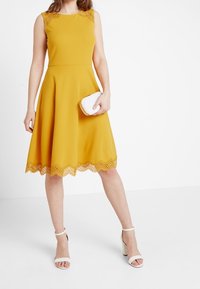 Světlé hořčicově žluté šaty bez rukávů s rozšířenou sukni, zdobené krajkovým lemem na spodním okraji. Vybavené bílou kabelkou a bílými sandály.